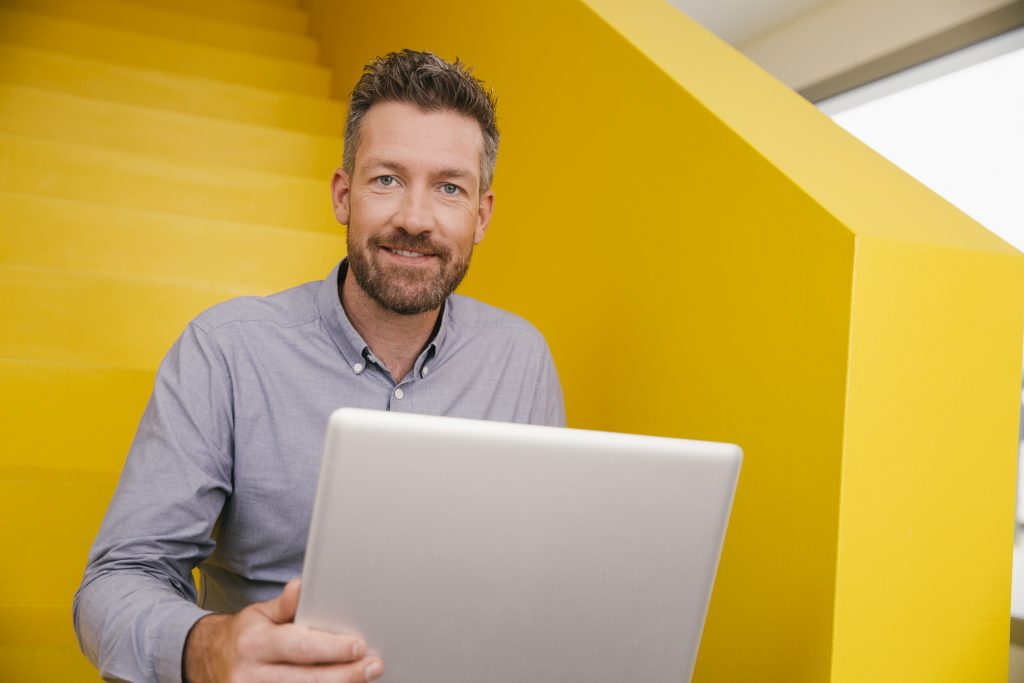 Portrait of smiling mature man with laptop sitting on yellow stairs